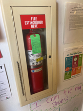 Fire extinguisher in lock box with glass cover. A chain with no attachment dangles from the box. Signage reads Fire Extinguisher Here.